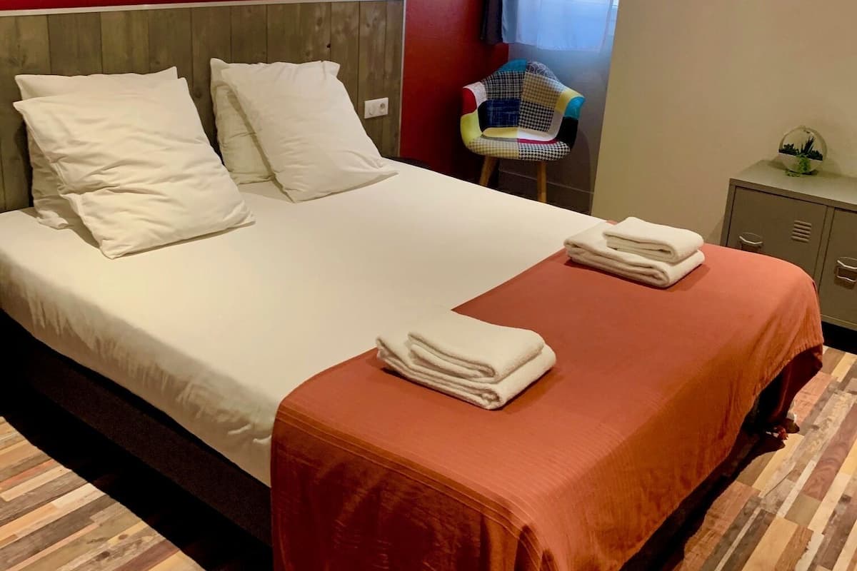 A neatly made bed with white linens and a brown throw blanket, a wooden headboard, a wall-mounted light, and a small table with a plant.