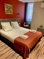 Una cama bien hecha con sábanas blancas y una manta marrón, cabecera de madera, una lámpara de pared y una mesita con una planta.