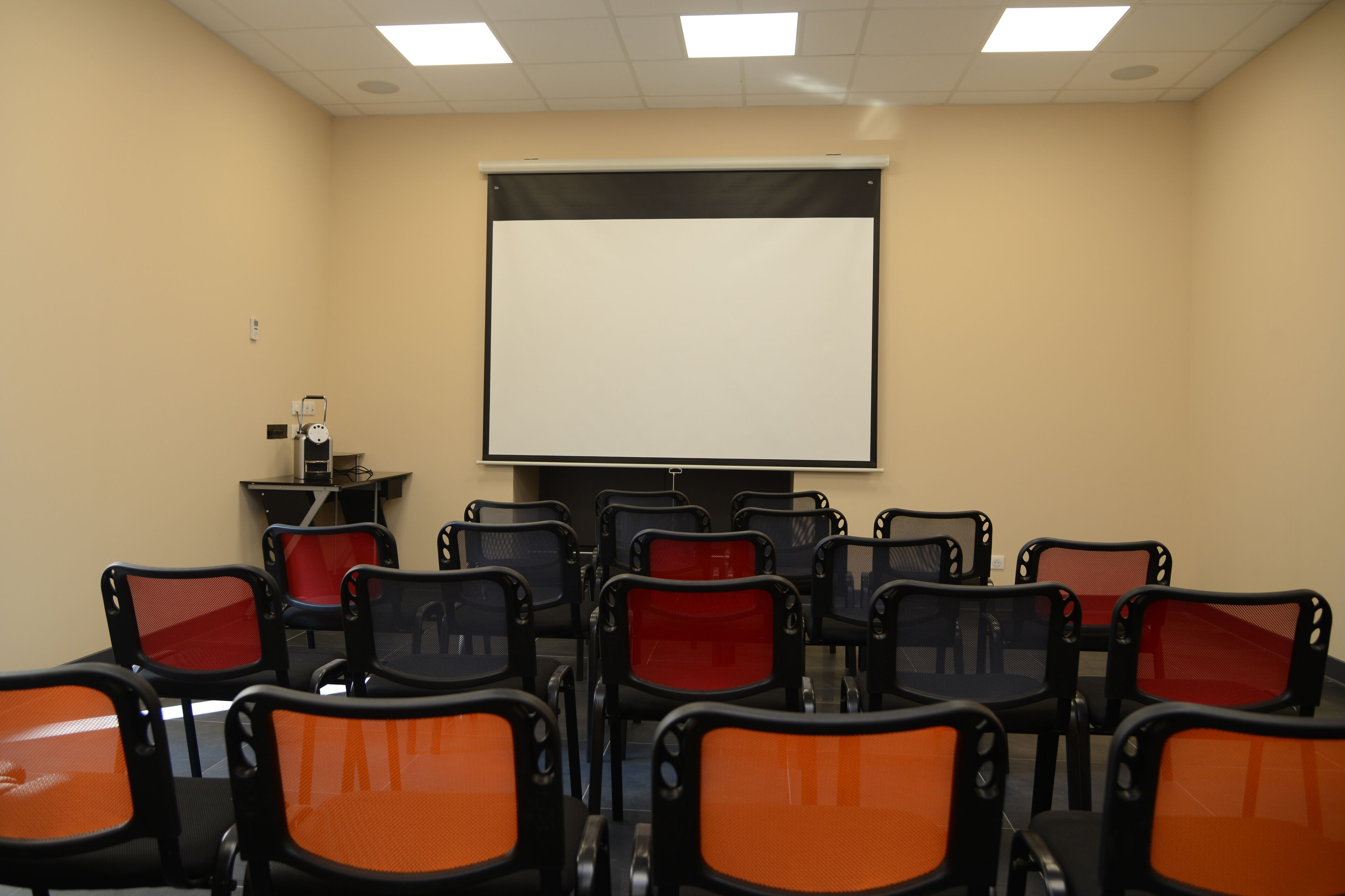 Une salle de conférence équipée d’un grand écran de projection, de plusieurs rangées de chaises et d’une petite table sur laquelle est posé un projecteur.