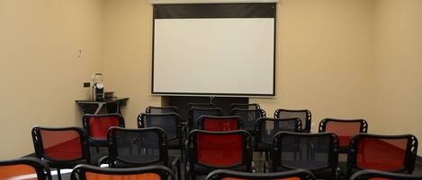 A conference room with a large projection screen, multiple rows of chairs, and a small table with a projector.