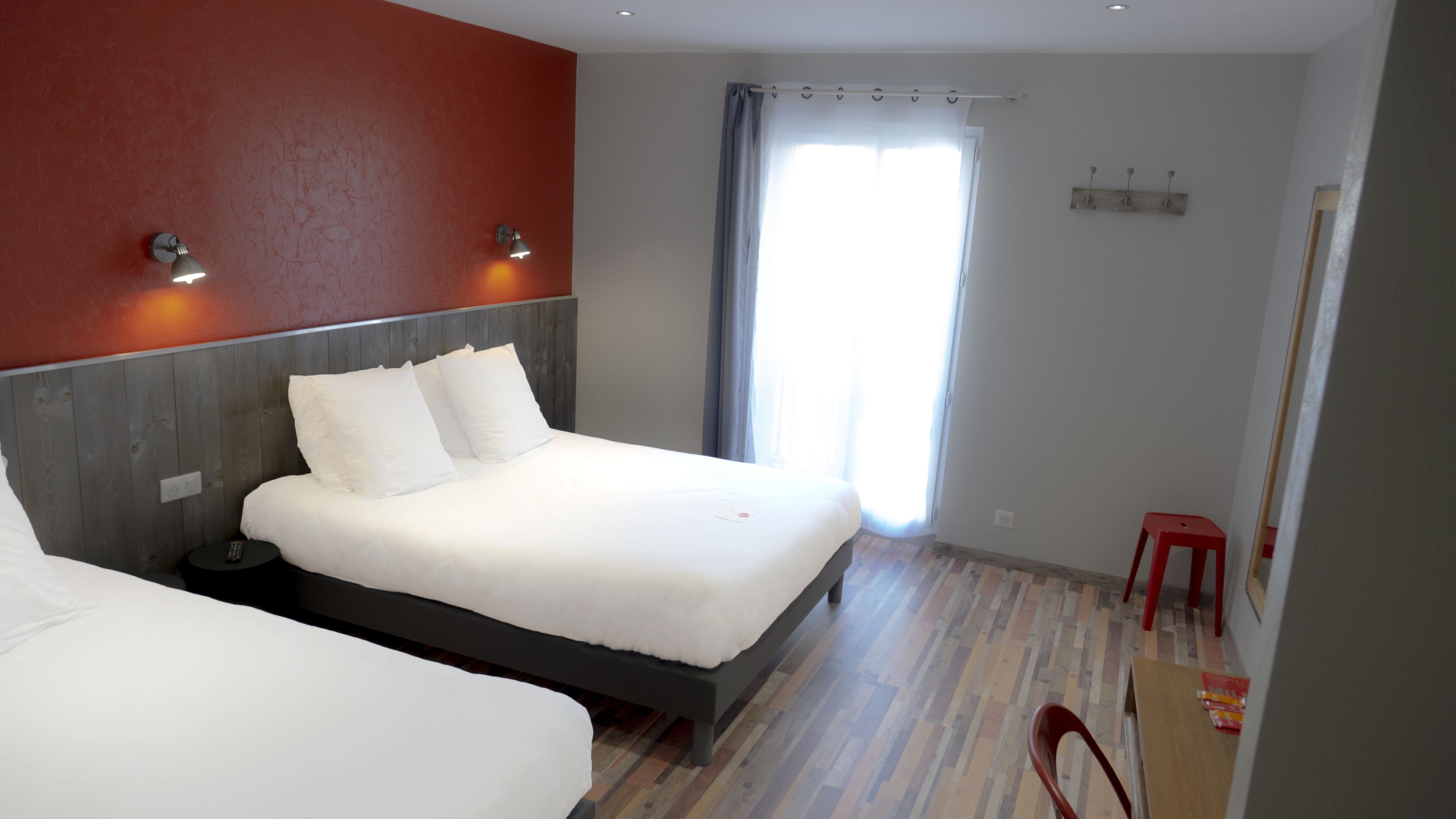 Une chambre d’hôtel avec un lit, une table de chevet, un mur rouge, une fenêtre avec des rideaux et une chaise rouge.