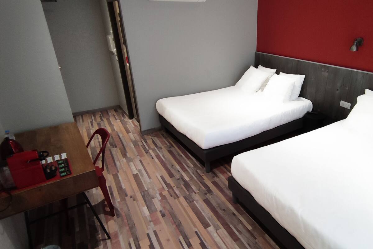 A hotel room with two single beds, a wooden floor, a red accent wall, and a small table with a red chair.