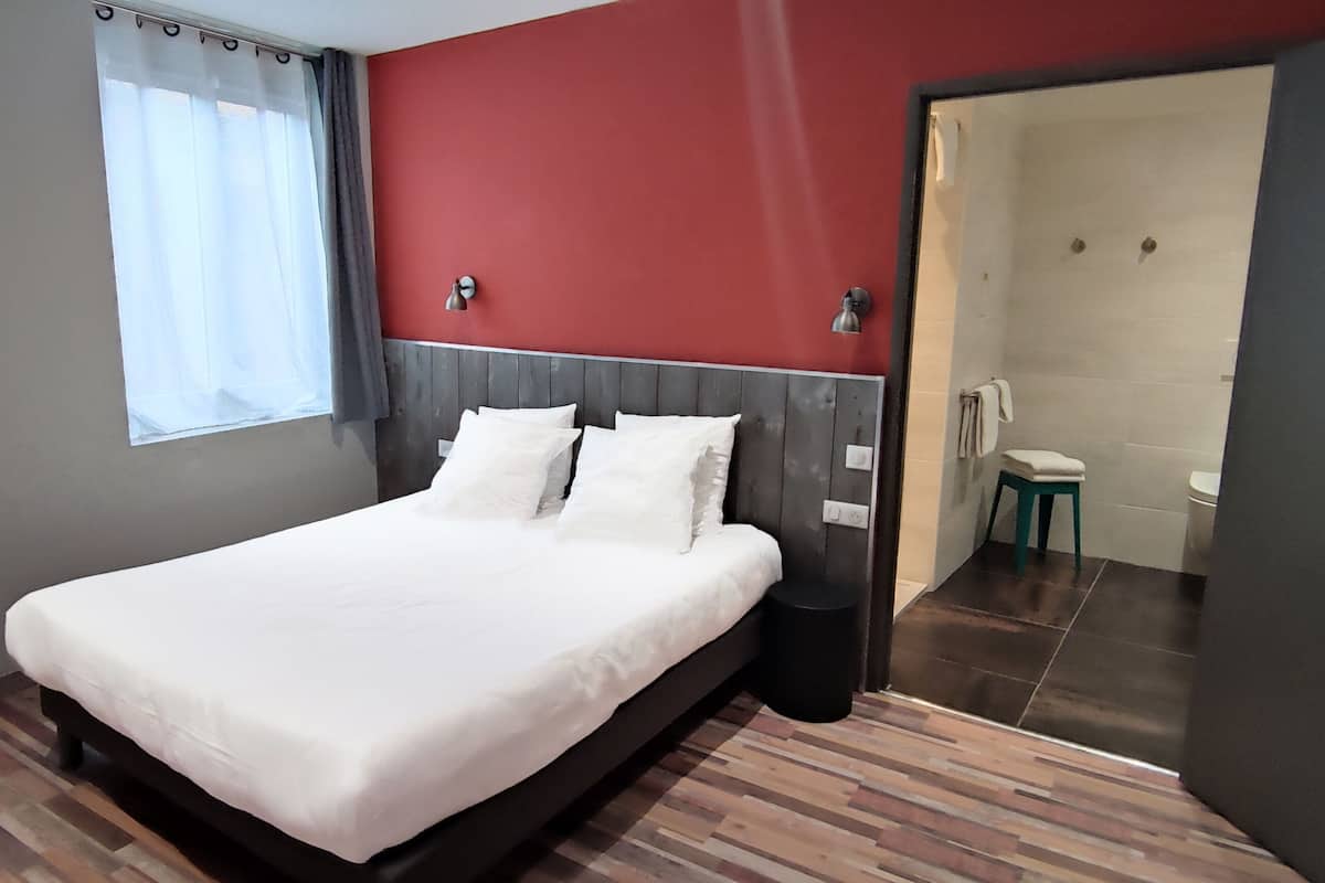 A modern hotel room with a large bed, a window with curtains, a red accent wall, and a visible bathroom.