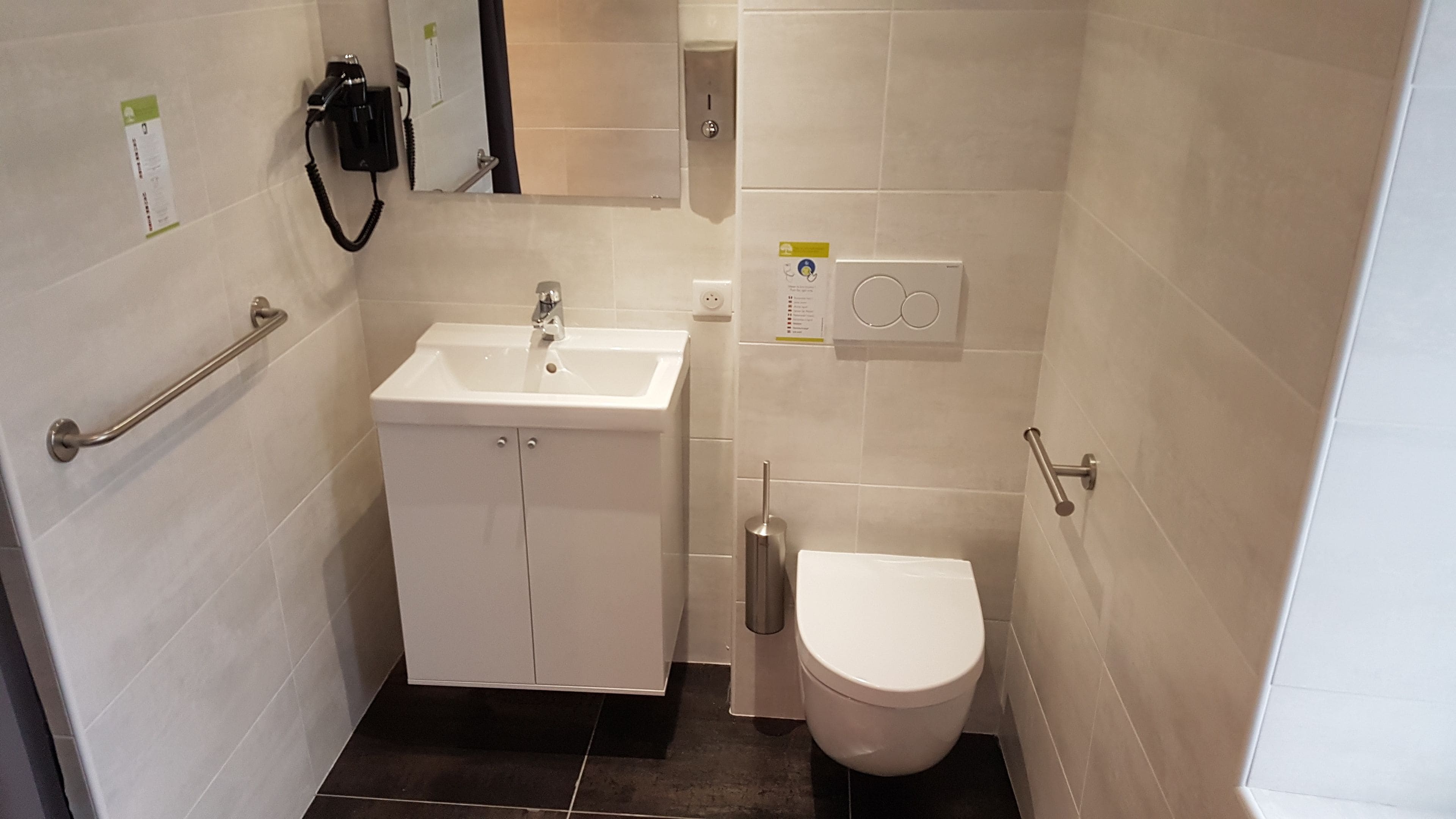 Une petite salle de bain moderne avec un lavabo blanc, des toilettes et un sèche-mains. Un porte-serviettes est fixé au mur et un miroir se trouve au-dessus du lavabo.