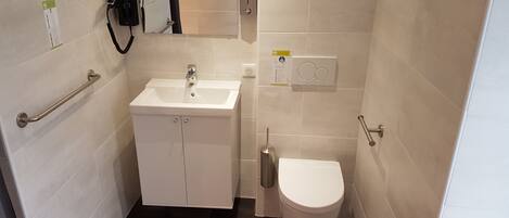 Une petite salle de bain moderne avec un lavabo blanc, des toilettes et un sèche-mains. Un porte-serviettes est fixé au mur et un miroir se trouve au-dessus du lavabo.