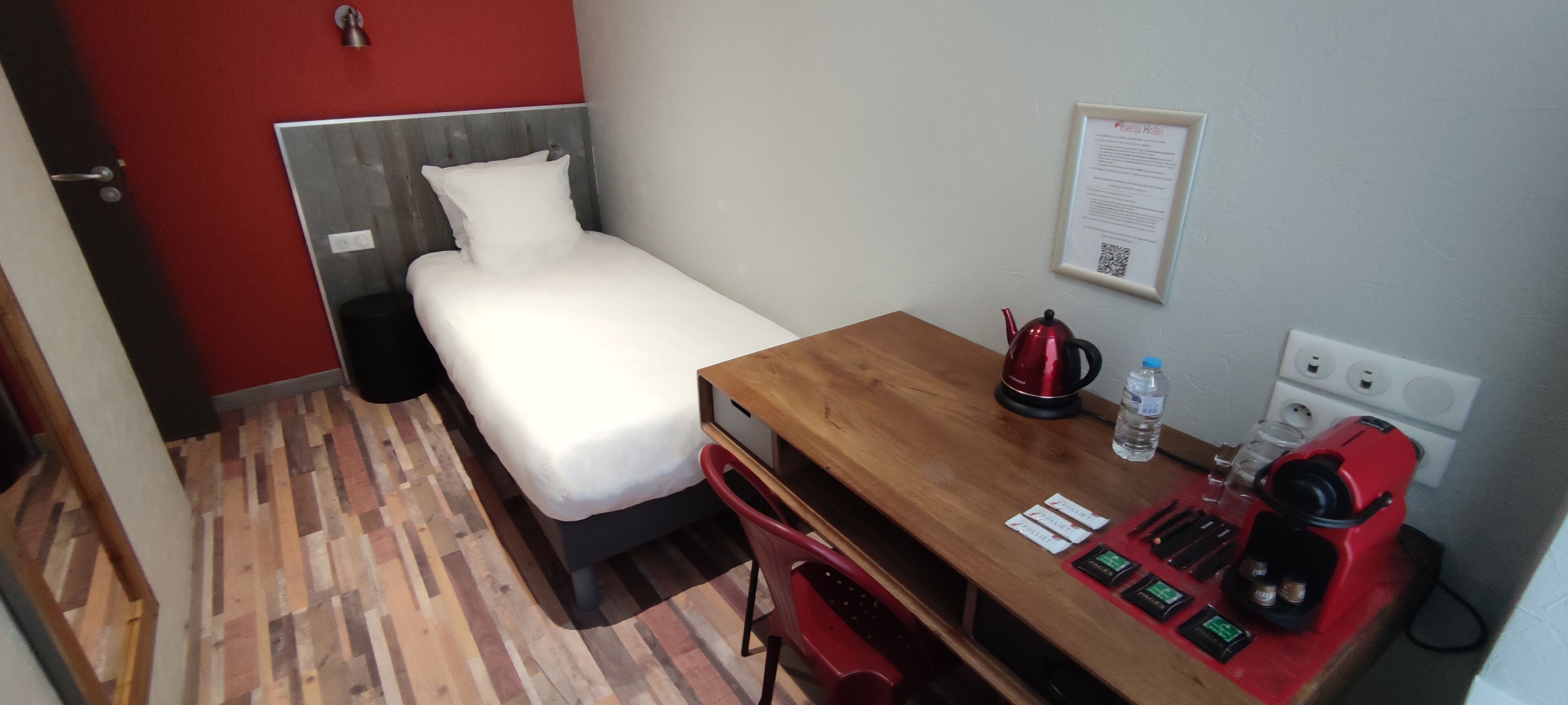 A small hotel room with a single bed, a wooden desk, a red coffee maker, a kettle, and a water bottle.