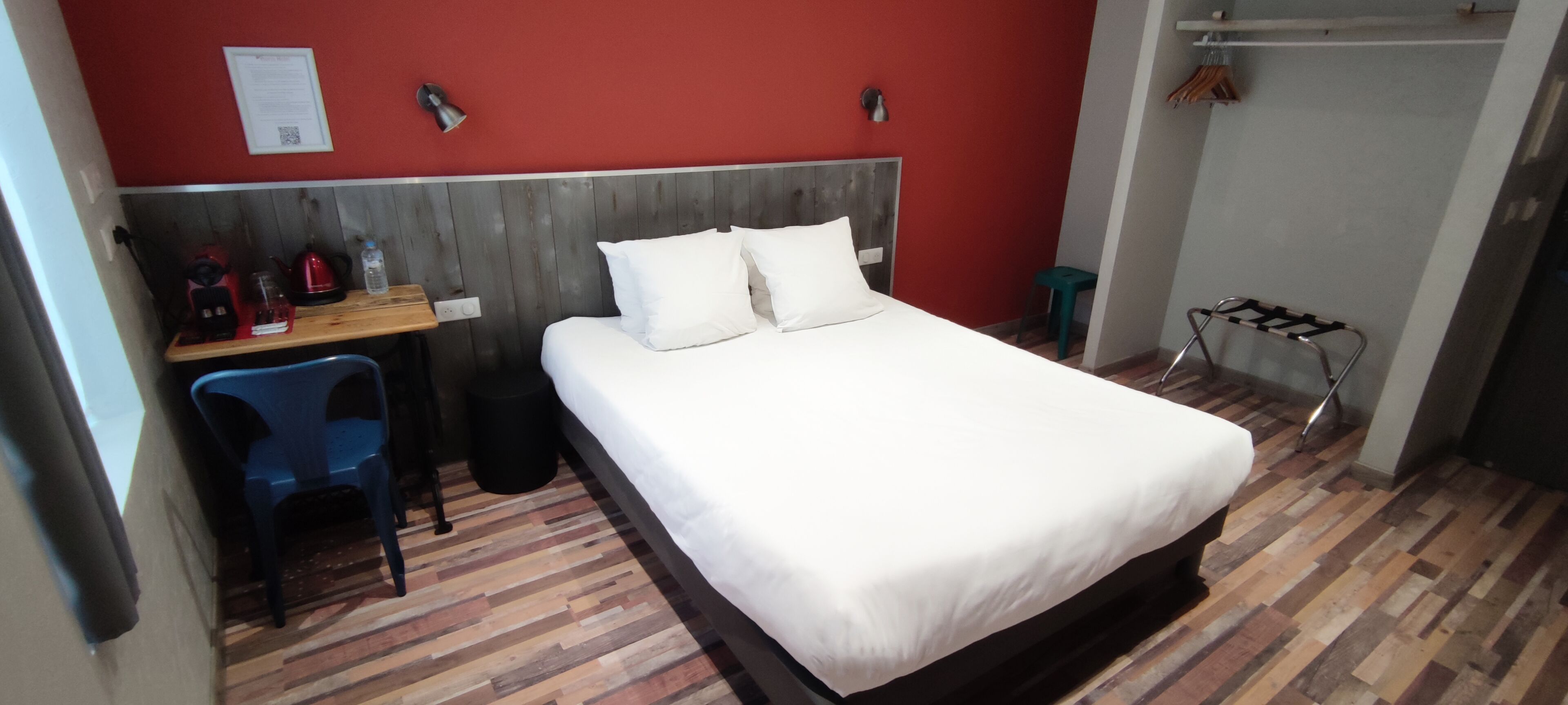 A hotel room with a bed, a desk, a chair, and a small stool.