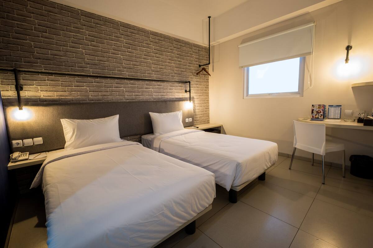Une chambre d’hôtel avec deux lits, un bureau, une chaise, un mur en briques et une fenêtre.