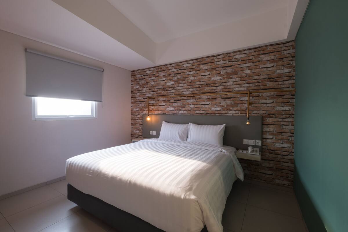Une chambre avec un mur décoré d’un motif en briques, un lit avec une literie blanche, une table de chevet avec une lampe et une fenêtre avec un store.
