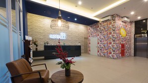 A hotel reception area with a modern design, featuring a reception desk, a seating area with a brown leather chair, a round wooden table with a vase of flowers, and a wall with a mosaic of colorful images. - Hotel 88 Bekasi By WH (Bekasi)