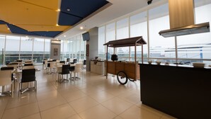 A modern dining area with tables and chairs, a central serving cart, and large windows offering views of the city. - Hotel 88 Bekasi By WH (Bekasi)