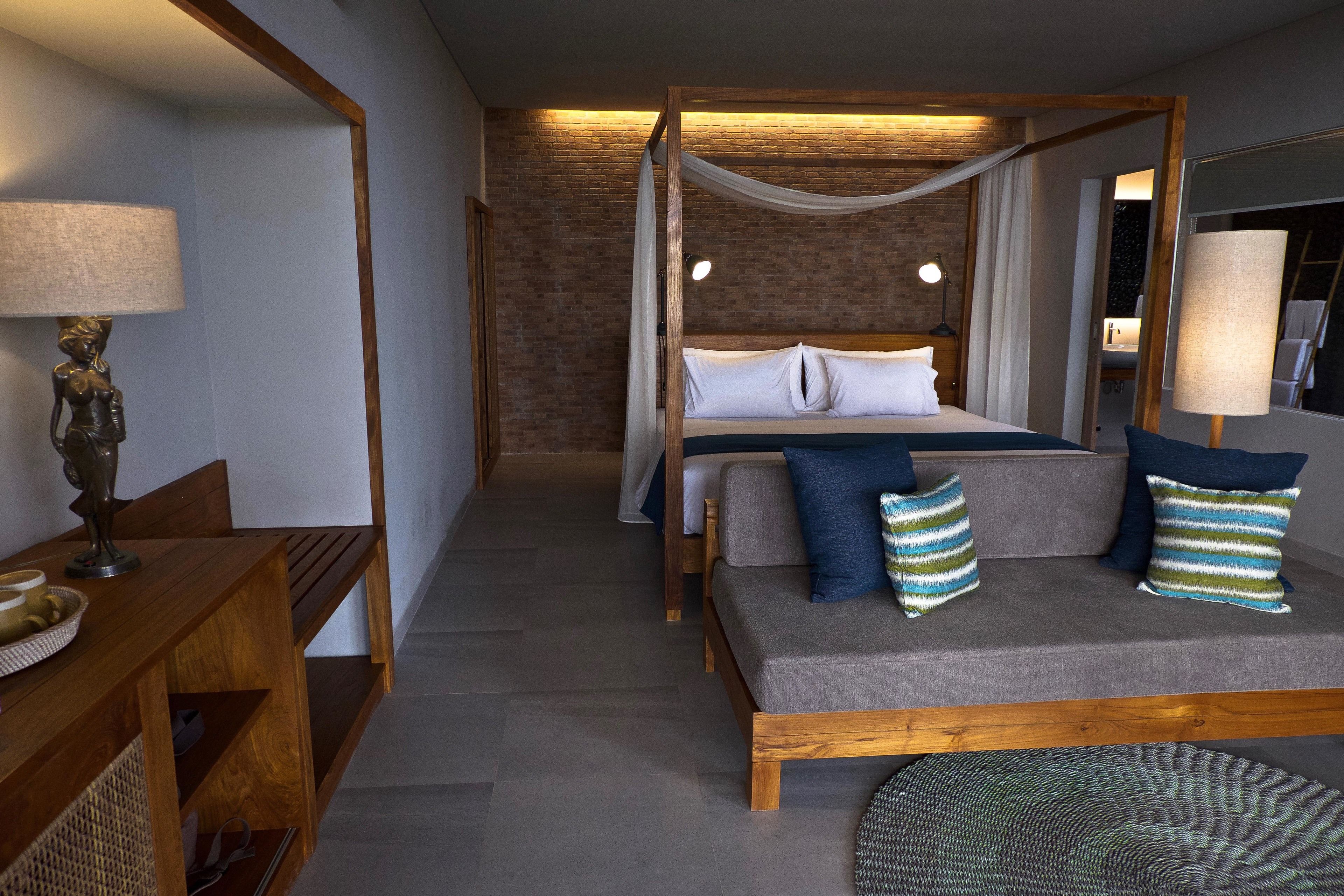 A bedroom with a four-poster bed, a sofa with cushions, a wooden desk with a lamp, and a round woven rug.
