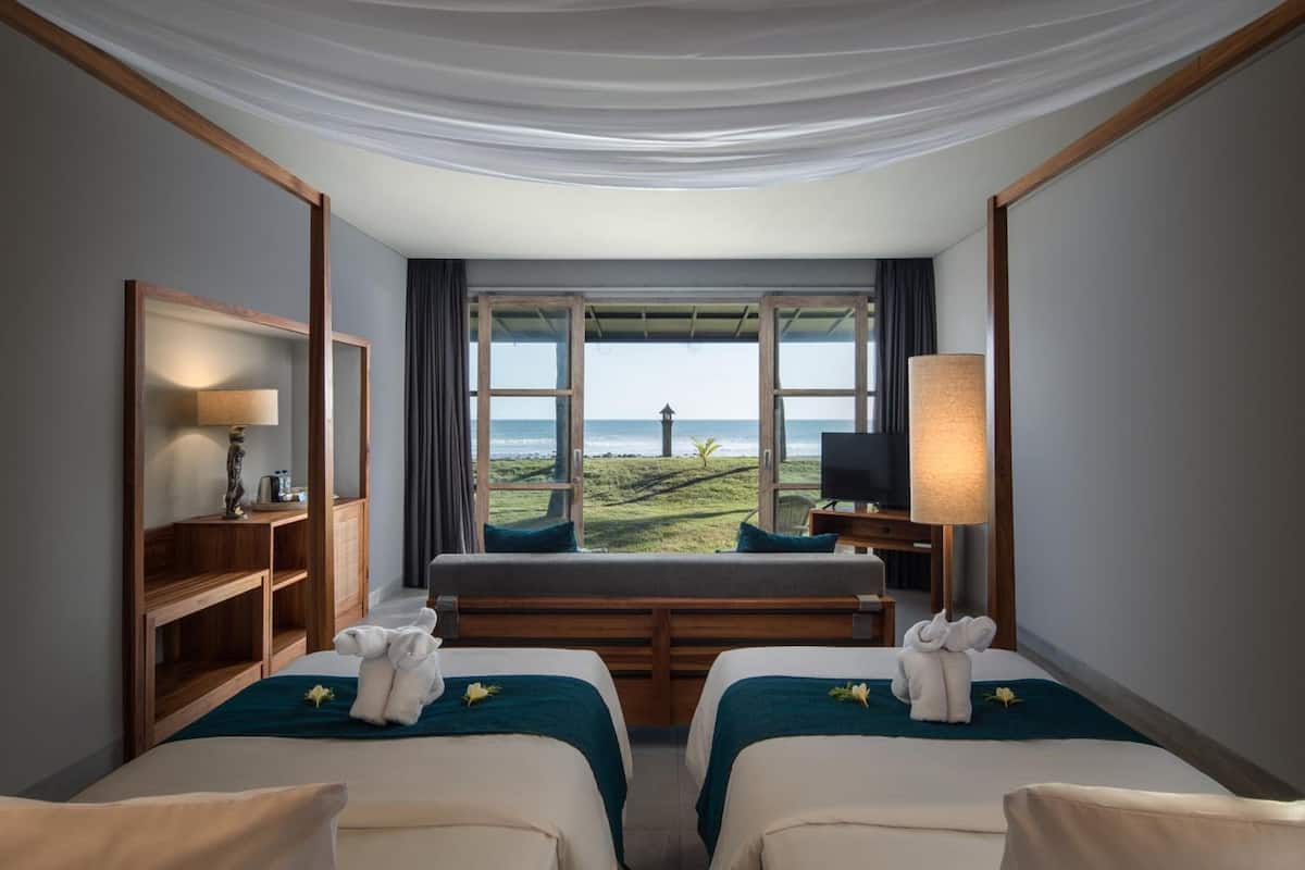 A hotel room with two beds, a TV, a desk, and a large window offering a view of the beach.