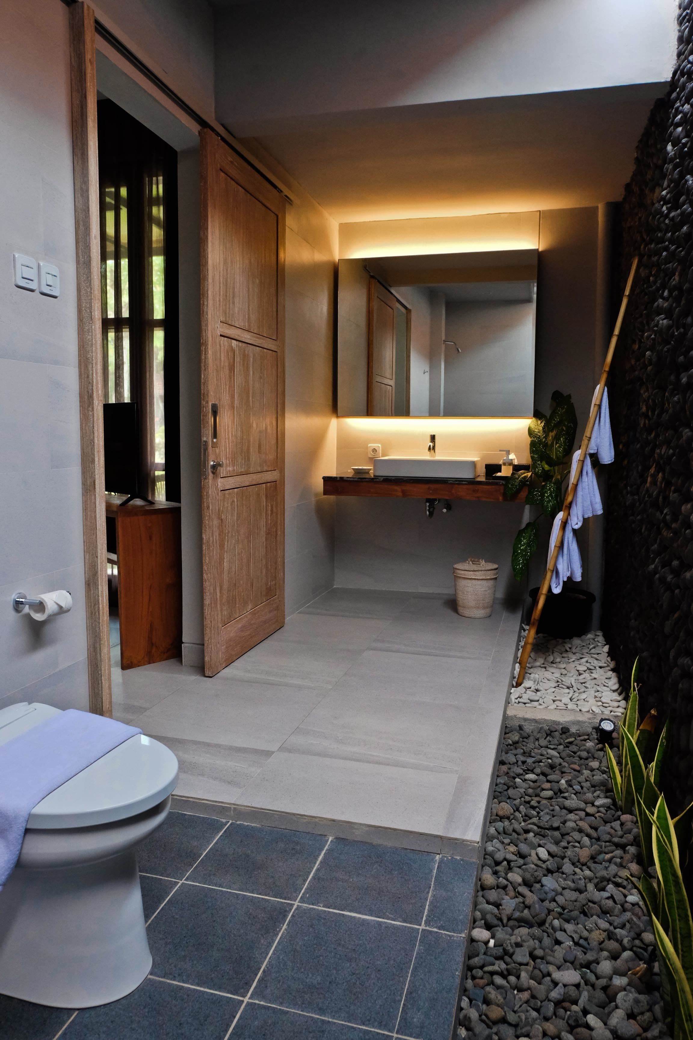 Een moderne badkamer met een houten deur, een witte wastafel en een spiegel erboven. Er is een toilet met een blauwe handdoek erop, en een klein plantje naast de wastafel.
