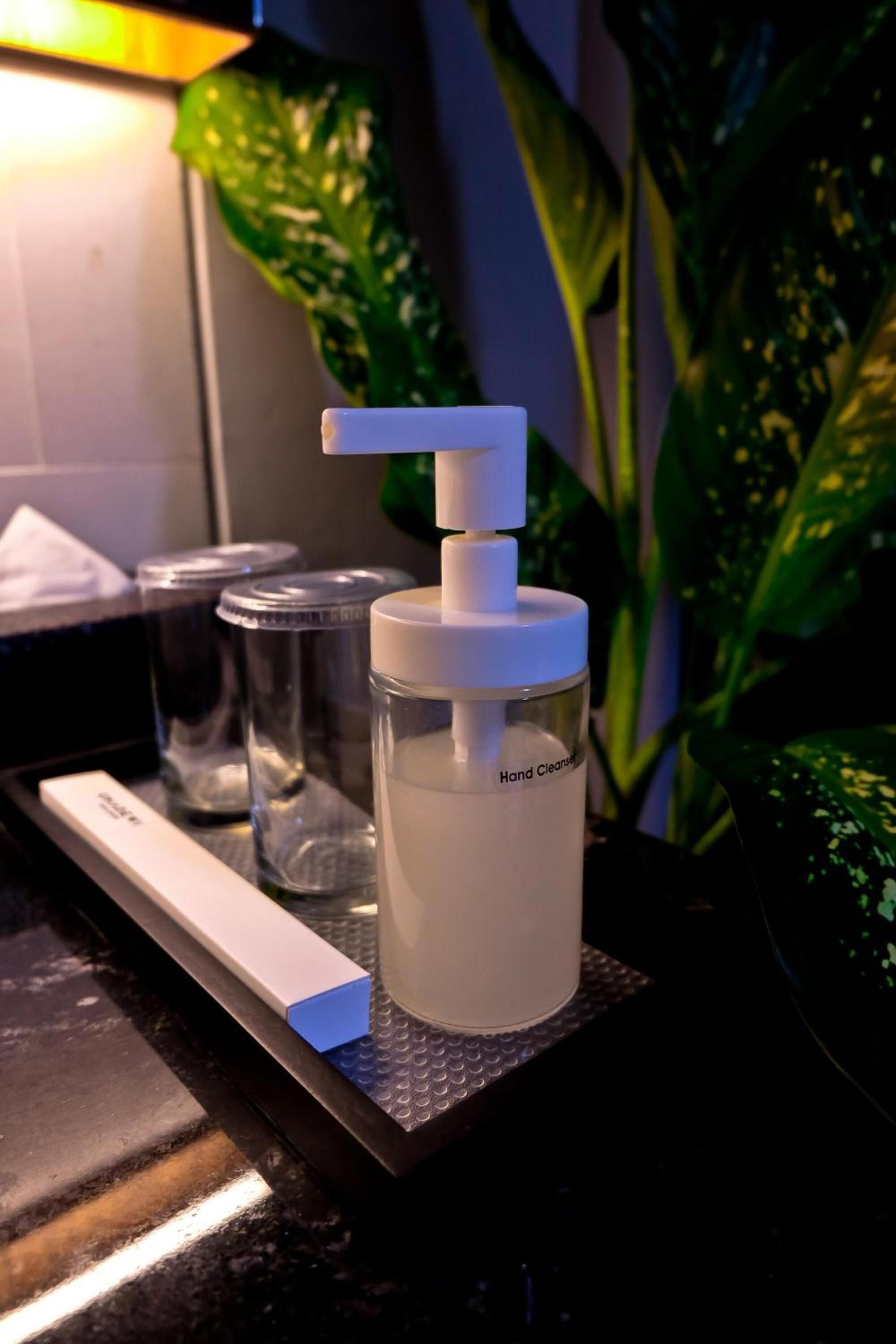 Un dispensador de gel antibacterial sobre una bandeja con etiqueta, recipientes de vidrio y una planta al fondo.