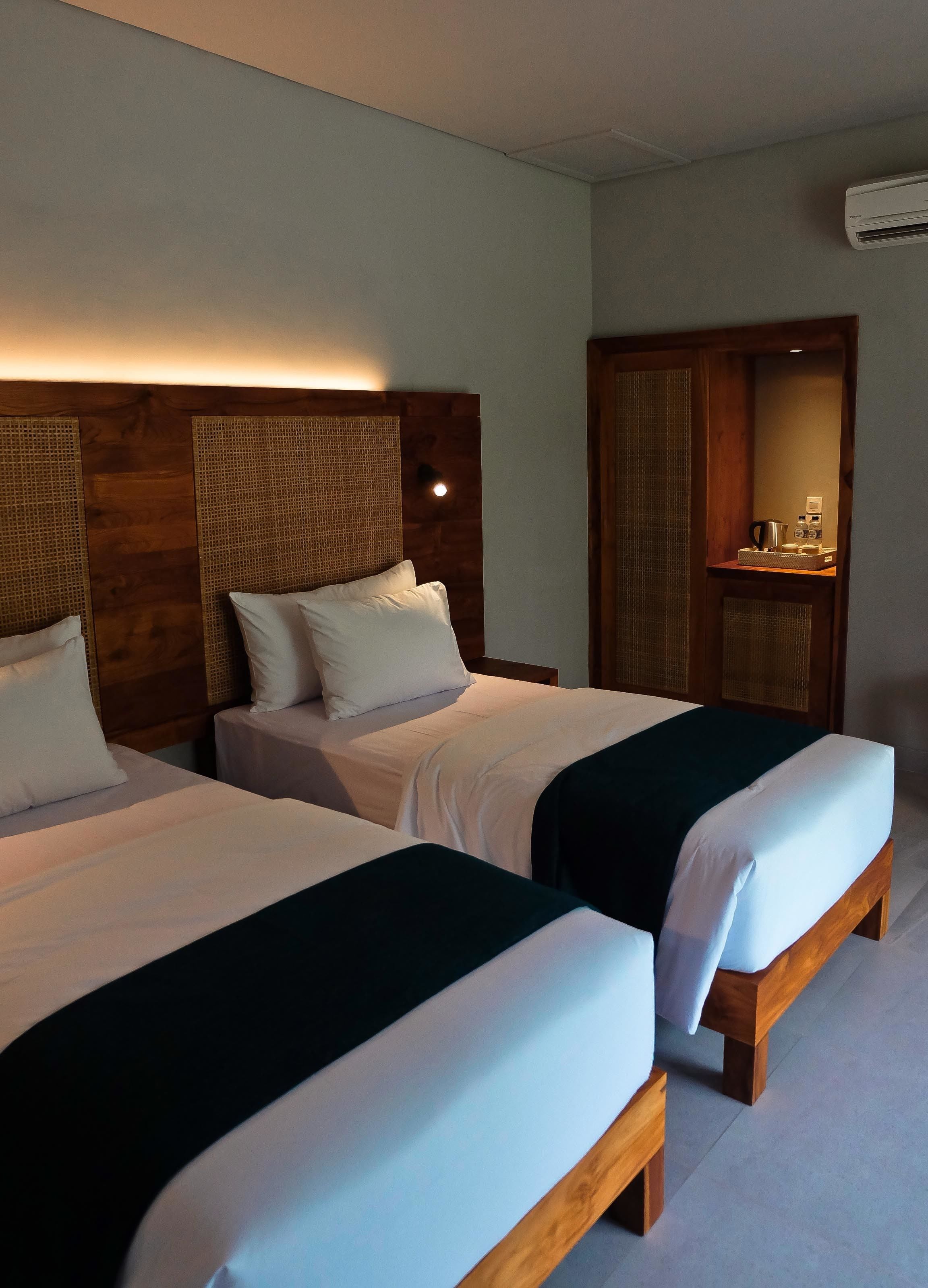 A hotel room with two beds, wooden headboards, a small table with a kettle, and a wall-mounted air conditioner.