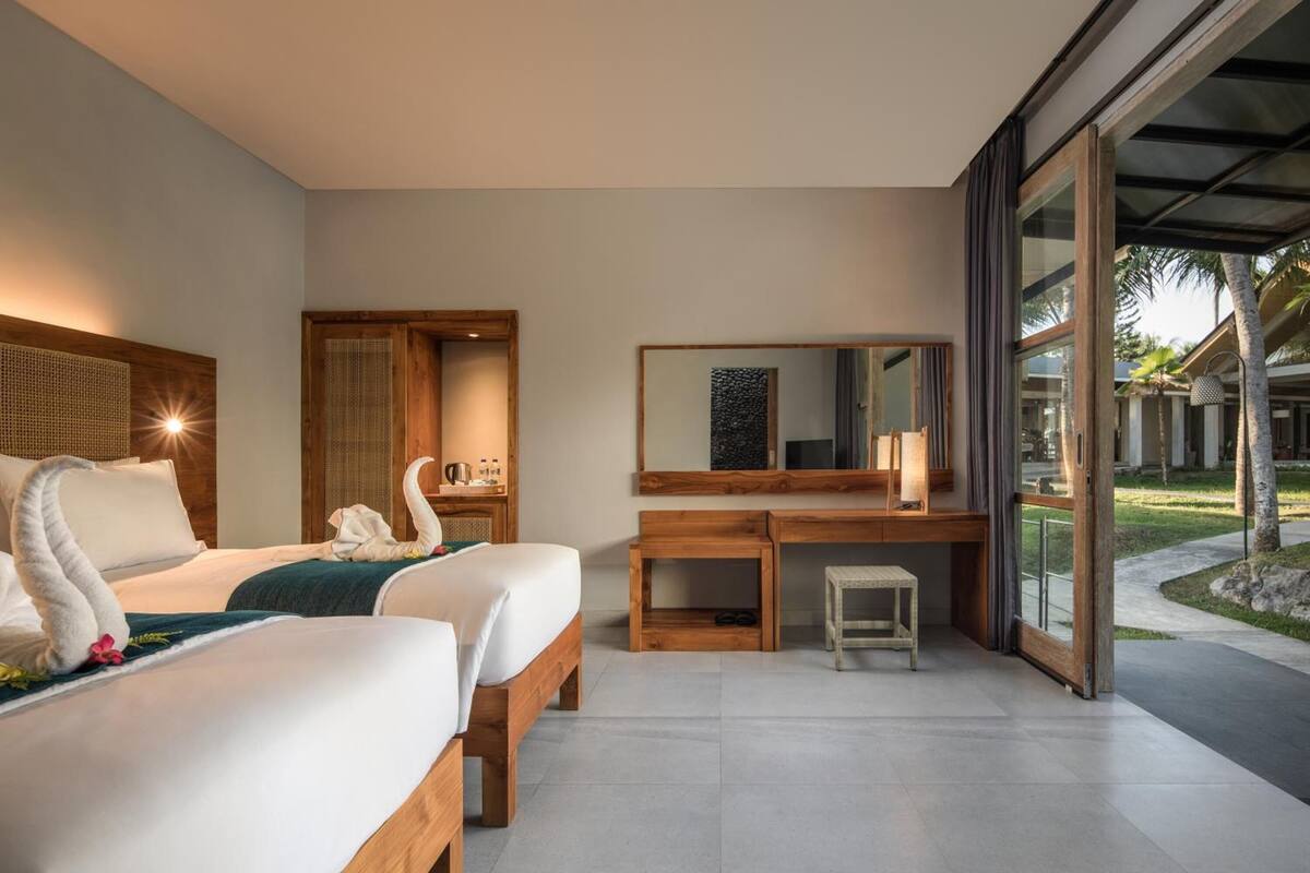 A modern hotel room with two beds, a wooden desk, a small stool, a large mirror, and a view of the outdoors.