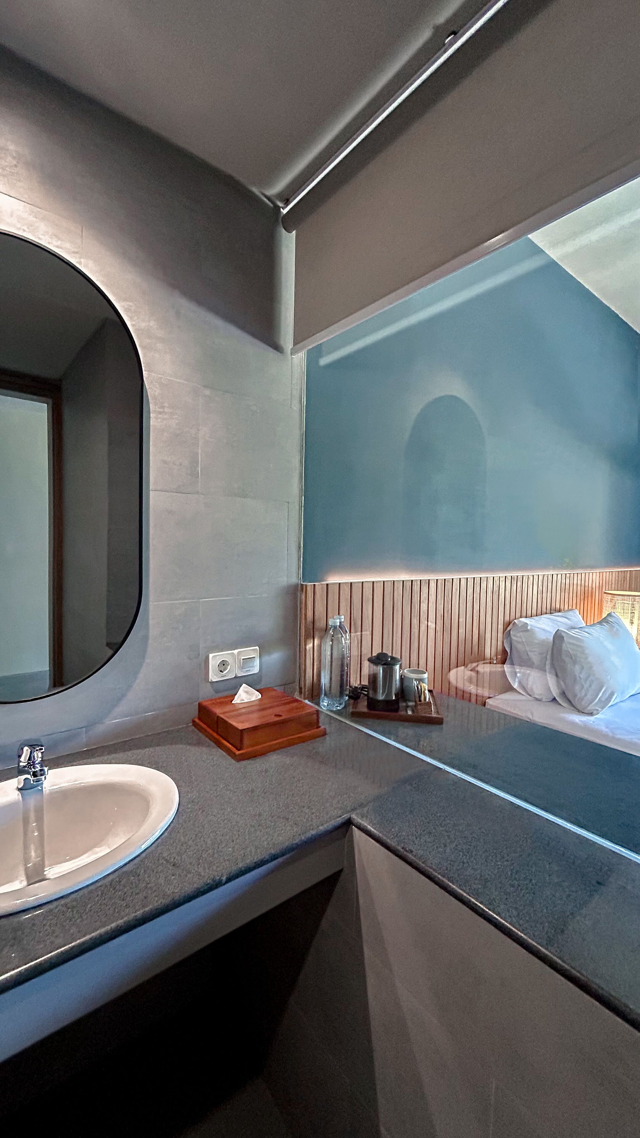 Un baño moderno con un espejo grande, un lavamanos y una mesita de noche con lámpara y botella.