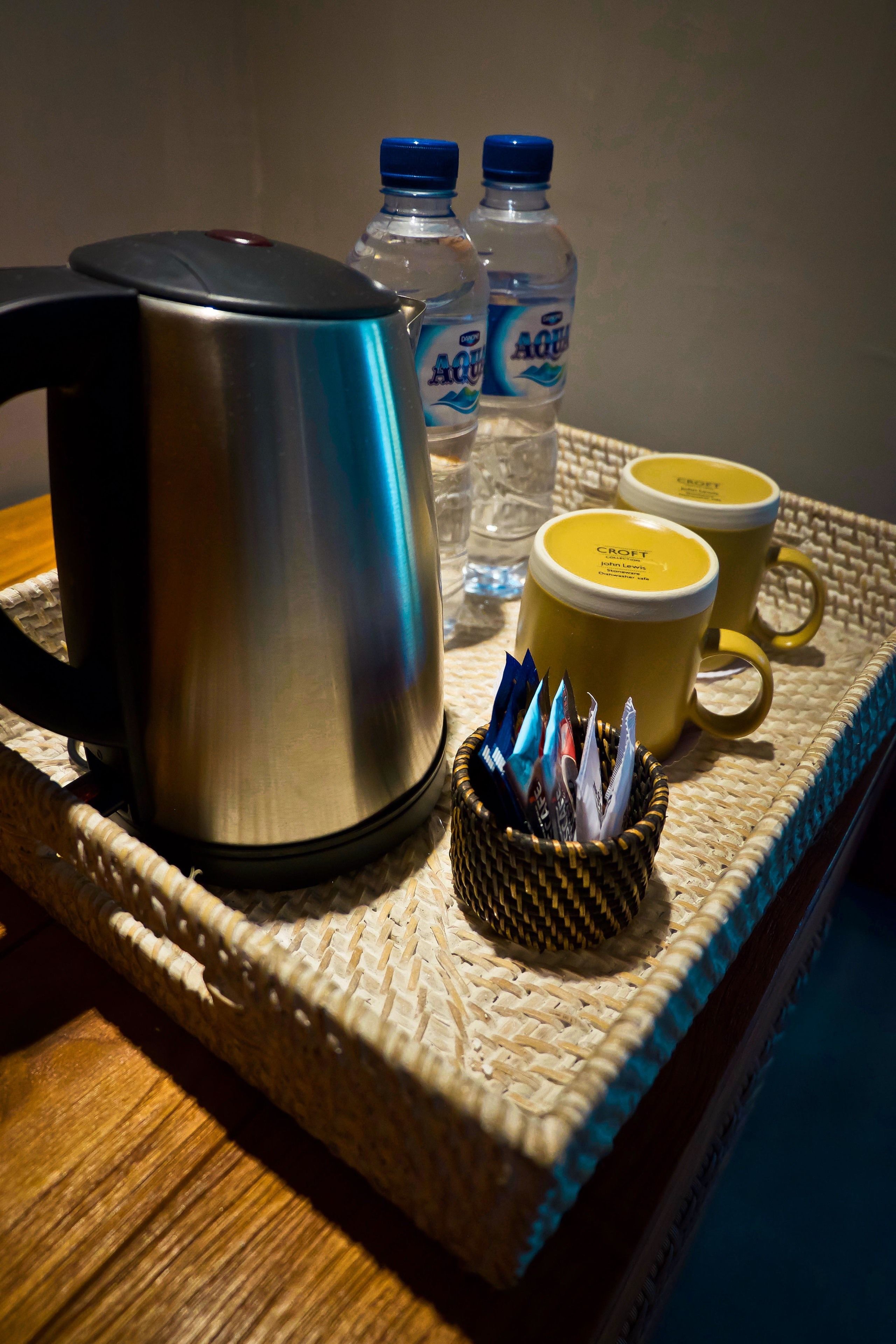 A stainless steel kettle, two water bottles, and two yellow mugs on a woven tray.