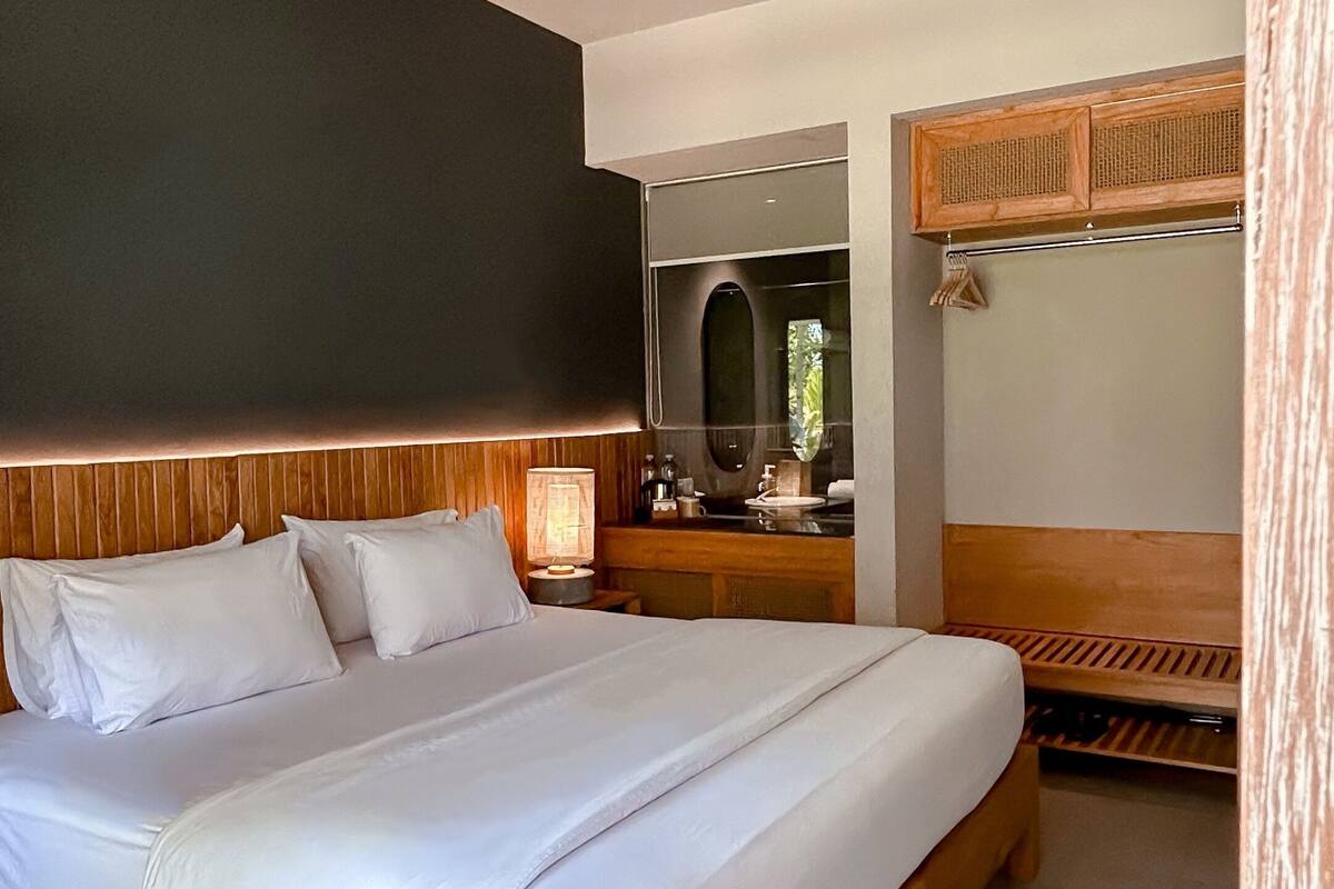 A modern bedroom with a large bed, a wooden headboard, a bedside table with a lamp, and a mirror above the sink.
