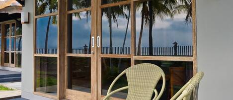 A covered outdoor seating area with a wicker chair and a glass door leading to a view of palm trees and a fence.