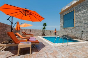 Ein Poolbereich mit Liegestühlen, orangefarbenen Sonnenschirmen und einer Steinmauer.