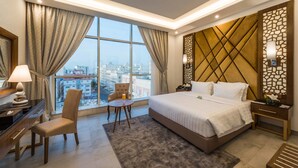A hotel room with a large bed, a desk, a chair, a small table, and a view of the city through large windows. - Boudl Quraish (Jeddah)