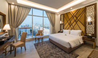 A hotel room with a large bed, a desk, a chair, a small table, and a view of the city through large windows.