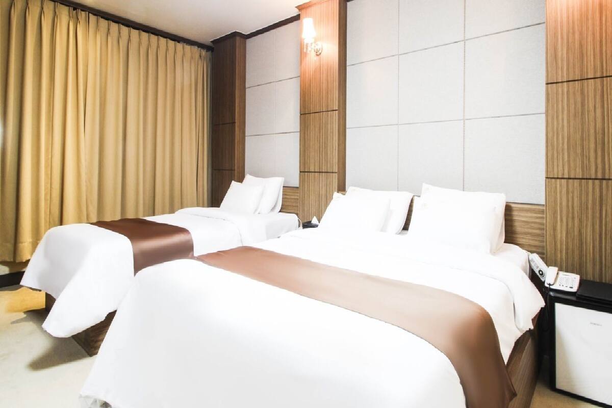 A hotel room with two beds, a nightstand, a lamp, and a large window with curtains.