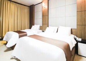Hotellihuone, jossa on kaksi sänkyä, yöpöytä, lamppu ja suuri ikkuna, jossa on verhot.