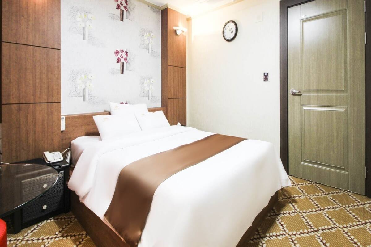 A hotel room with a bed, a bedside table, a telephone, and a wall clock.