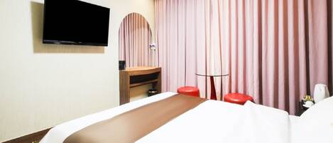 Hotellihuone, jossa on sänky, seinälle asennettu taulutelevisio, puinen yöpöytä, pieni pyöreä pöytä ja punainen jakkara.