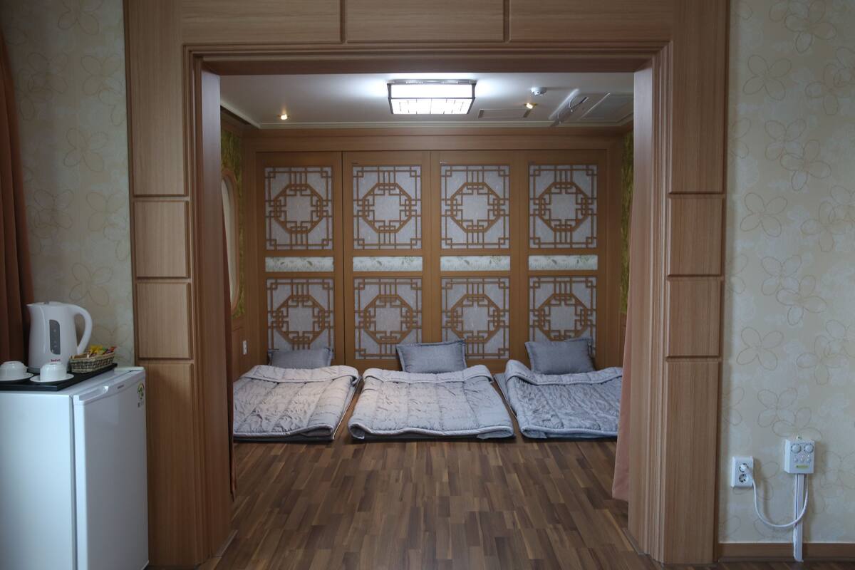 A room with a wooden floor, a bed with a grey blanket, a white kettle on a small table, and a wall with a geometric pattern.