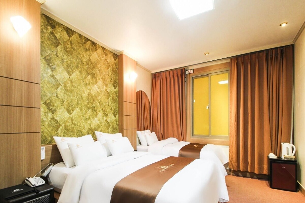 A hotel room with two beds, a minibar, a telephone, a window with curtains, and a wall with a textured pattern.