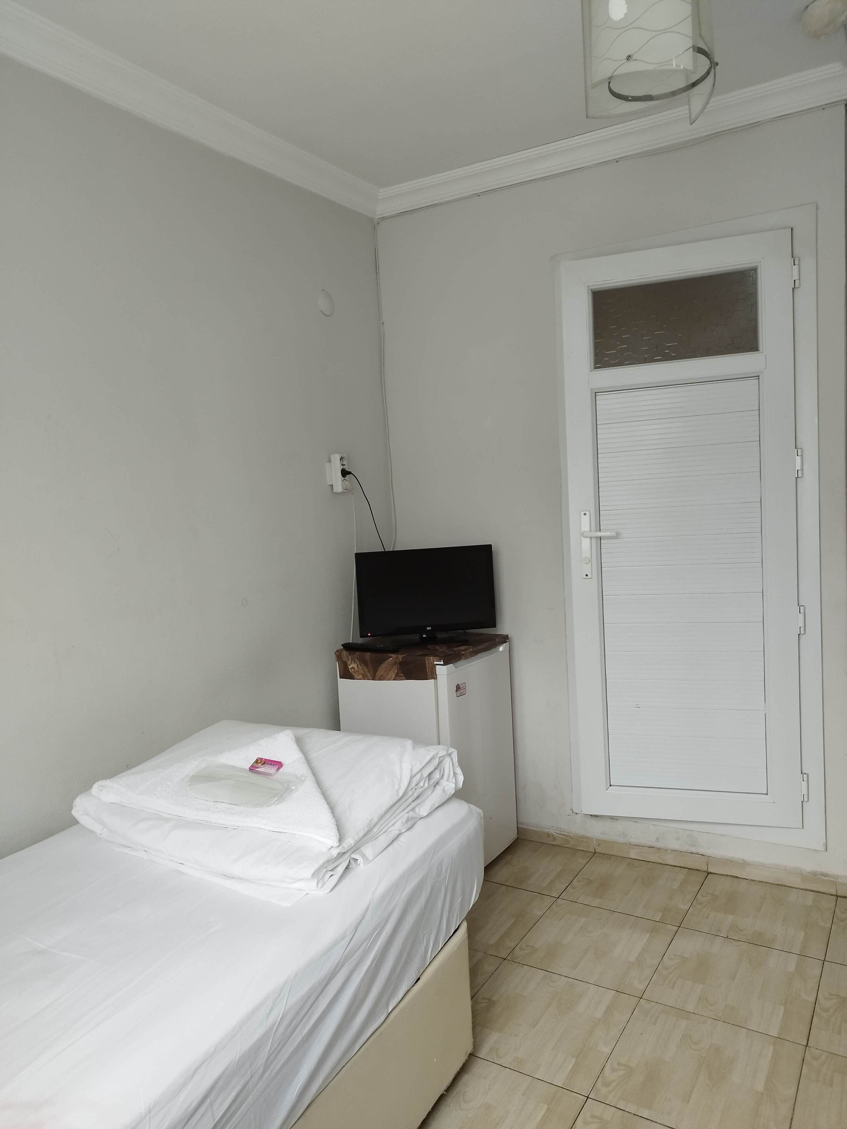 Una habitación pequeña y limpia, con una cama, un televisor sobre un soporte de madera y una puerta blanca.