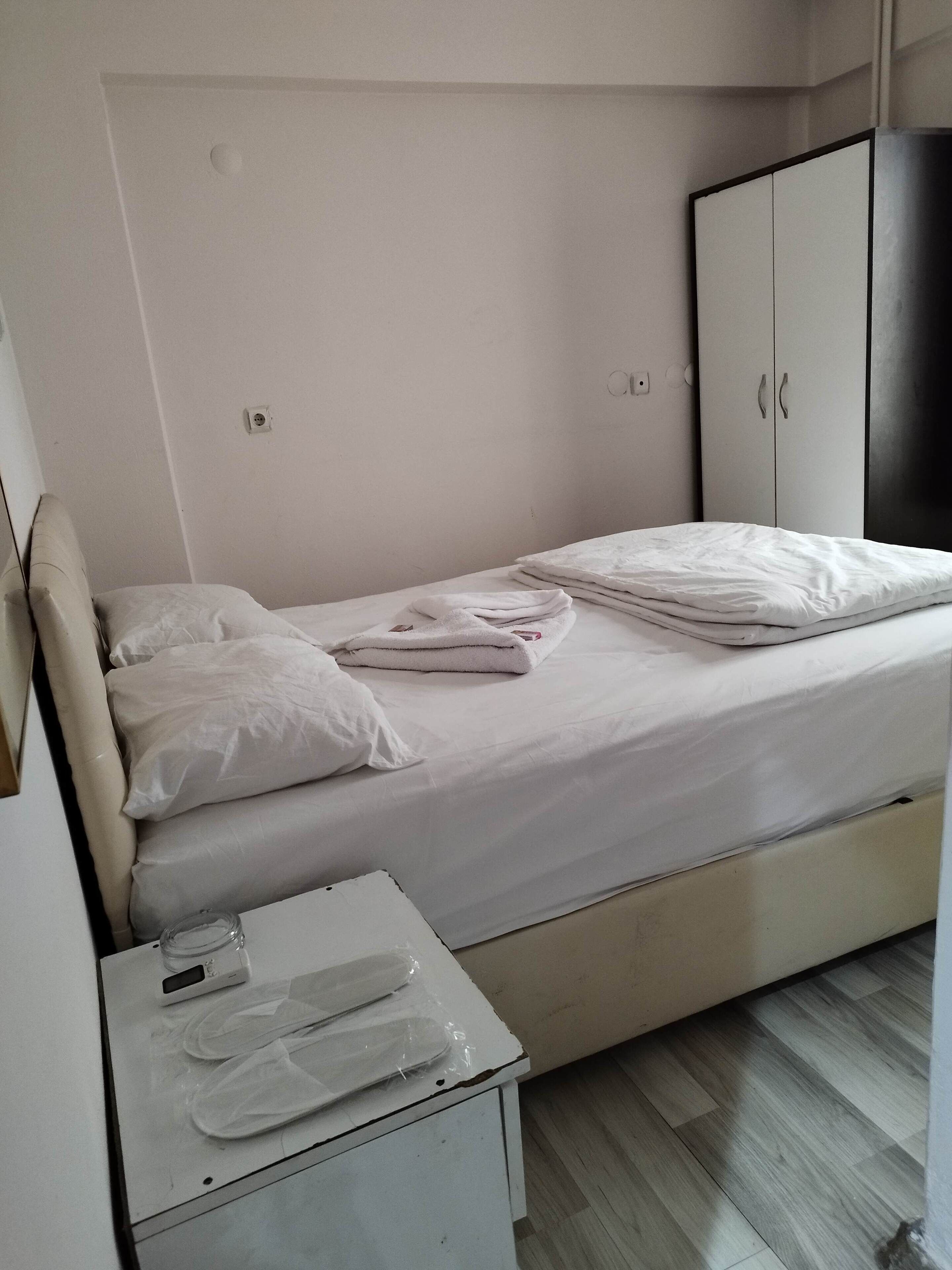 A small, neatly made bed with white linens, a bedside table with a digital device and a plastic bag, and a wardrobe with a dark door.