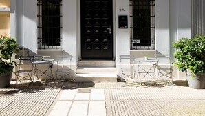 Un edificio de dos pisos con una puerta negra, un balcón con barandillas de hierro forjado y un pequeño espacio al aire libre con sillas y mesas.
