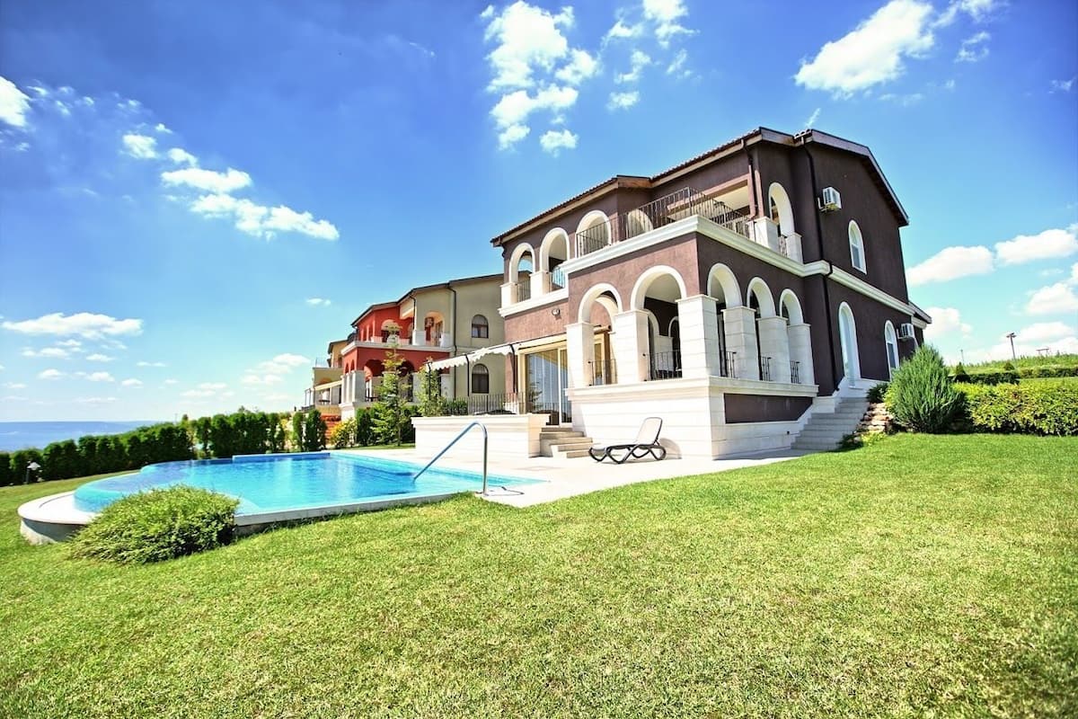 A large, multi-story house with a swimming pool, a well-maintained lawn, and a clear view of the sea.