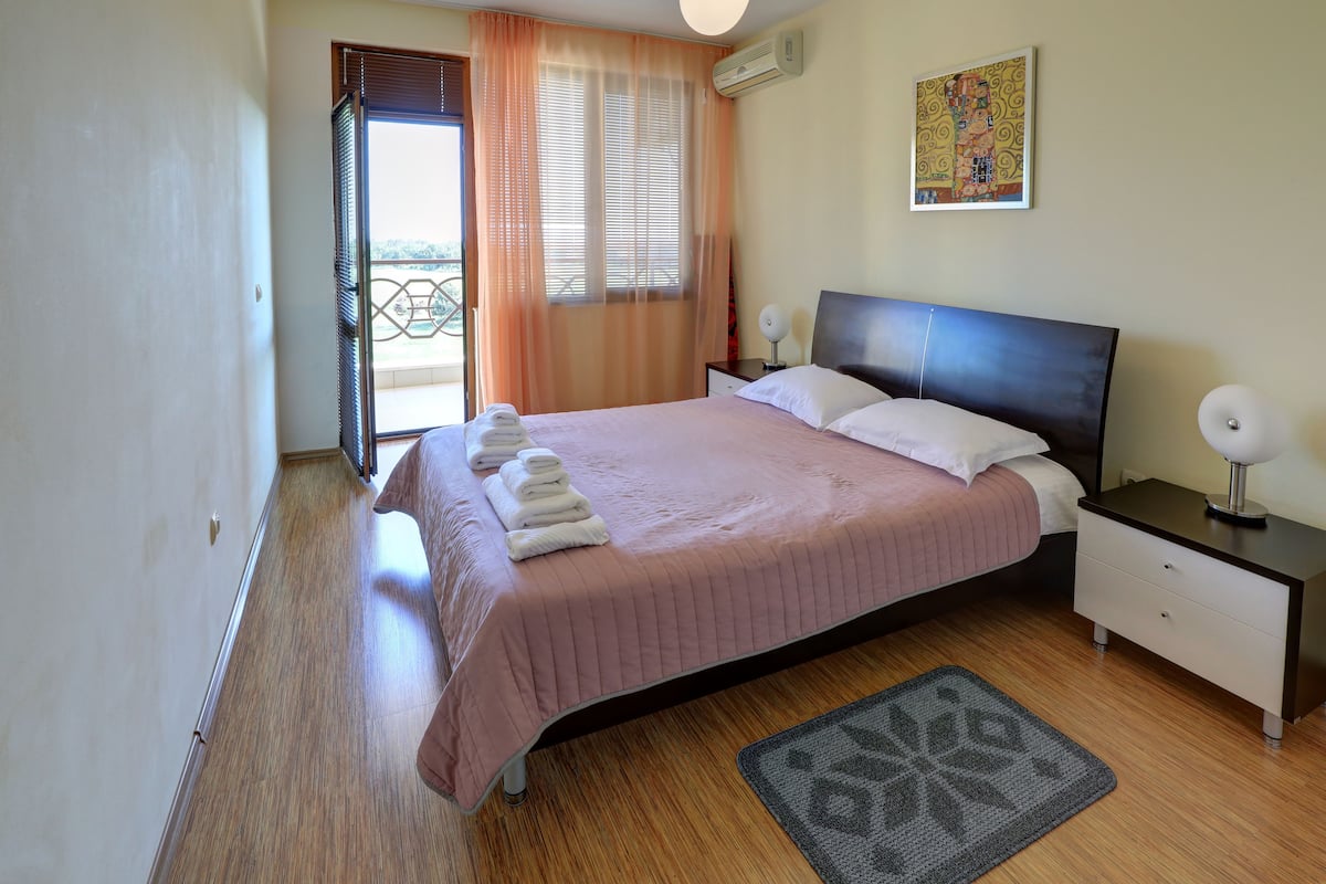 A bedroom with a bed, bedside tables, a nightstand, a window with curtains, a door, and a wall-mounted air conditioner.