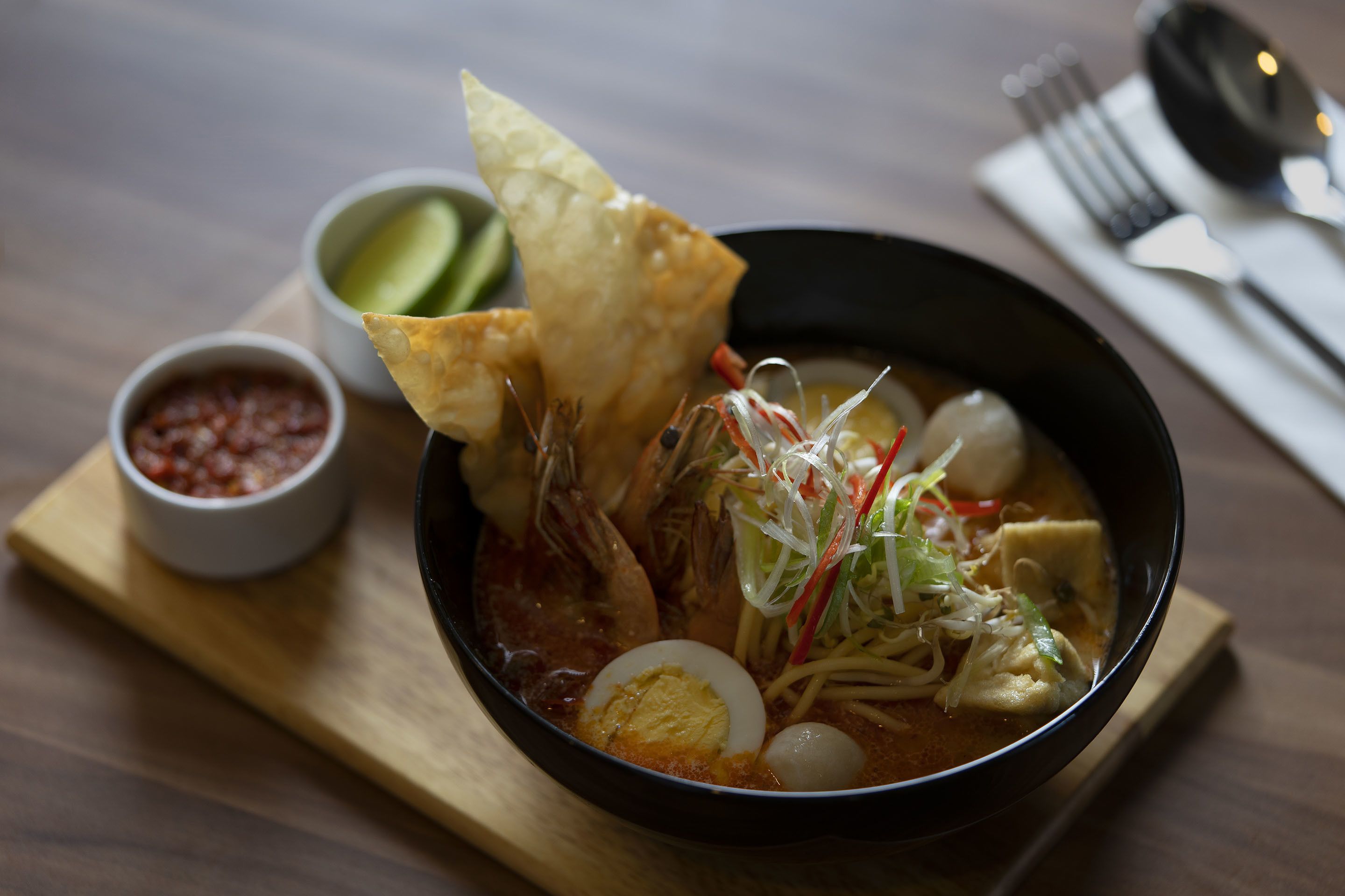 A bowl of soup with various ingredients, a piece of bread, and a small bowl of sauce on a wooden board.