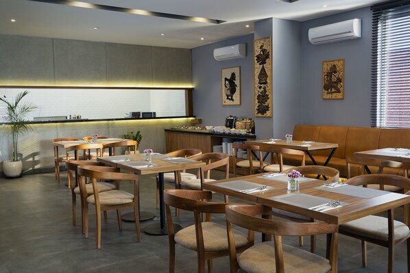 A modern dining area with wooden tables and chairs, a kitchen in the background, and decorative wall art.