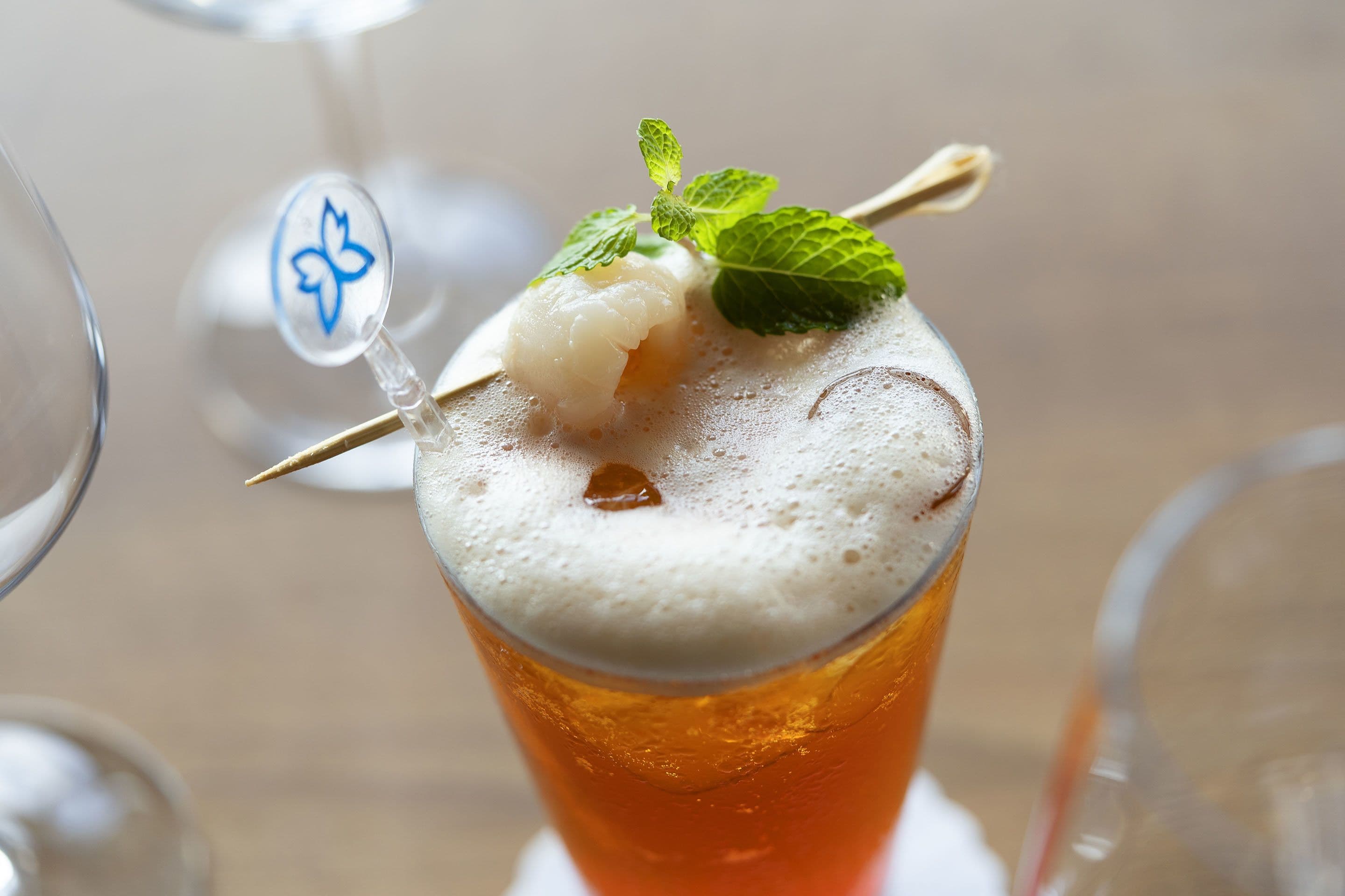 Ein Glas Bier mit Schaumkrone und einer Minzblatt-Garnierung, ein blaues Logo auf dem Glas.