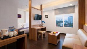 A modern hotel room with a wooden desk, a comfortable sofa, a television, and a large window offering a city view.