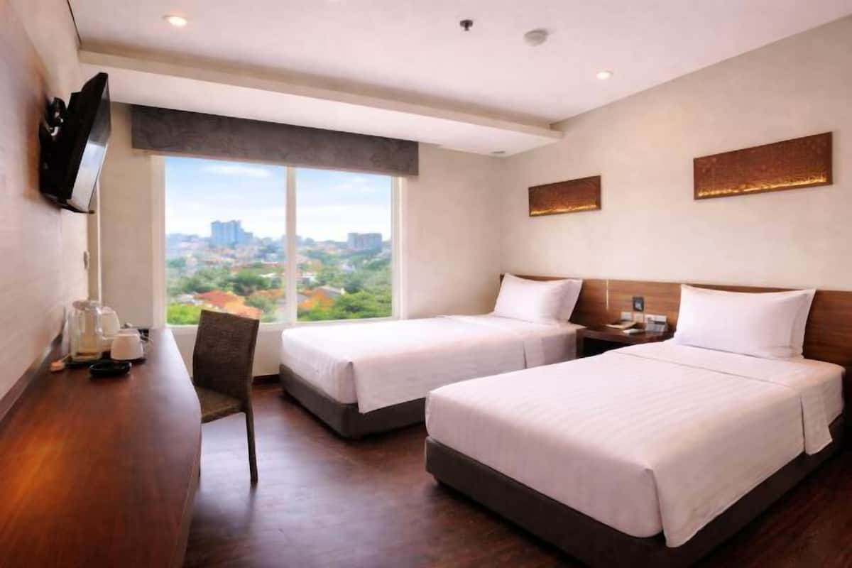 Une chambre d’hôtel avec deux lits, un téléviseur fixé au mur, une grande fenêtre donnant sur la ville et un bureau en bois avec une chaise.