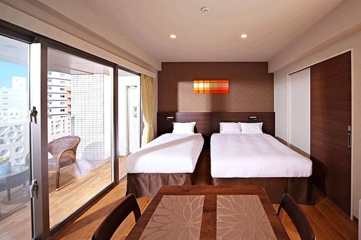 Ein Hotelzimmer mit zwei Betten, einem Esstisch und Blick auf die Stadt durch eine Schiebetür.