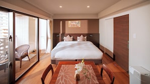 A hotel room with a large bed, a dining table with chairs, a wicker chair, and a small table with a vase of flowers. - SmartCondo Tomari (Naha)