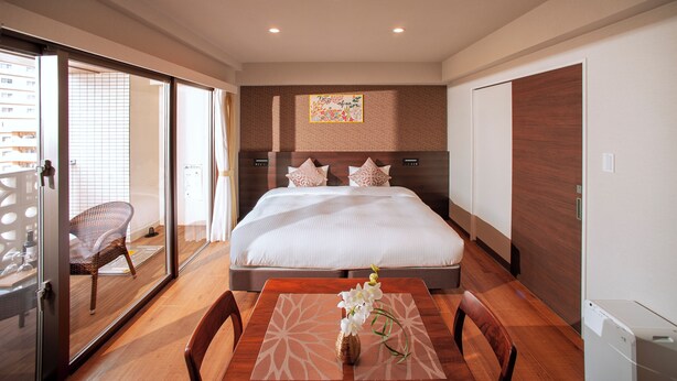 A hotel room with a large bed, a dining table with chairs, a wicker chair, and a small table with a vase of flowers.