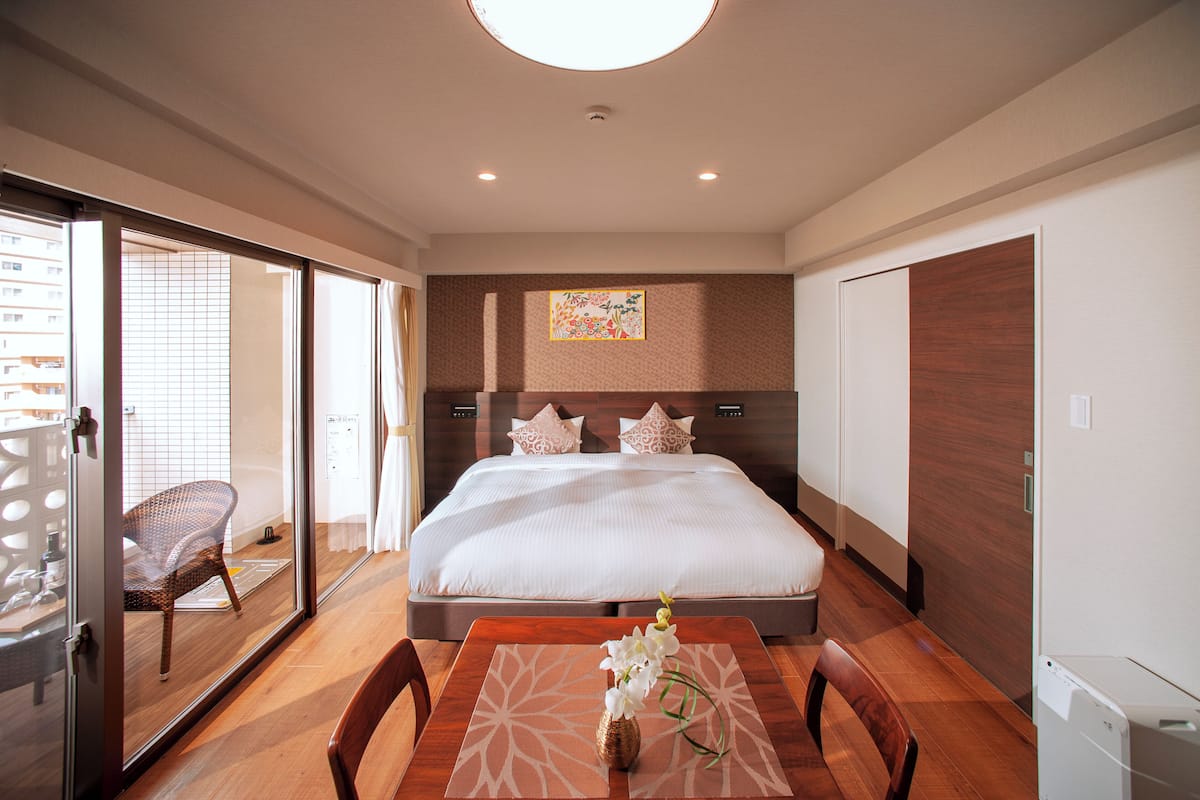 Ein Hotelzimmer mit einem großen Bett, einem Esstisch mit Stühlen, einem Korbsessel und einem kleinen Tisch mit Blumen.
