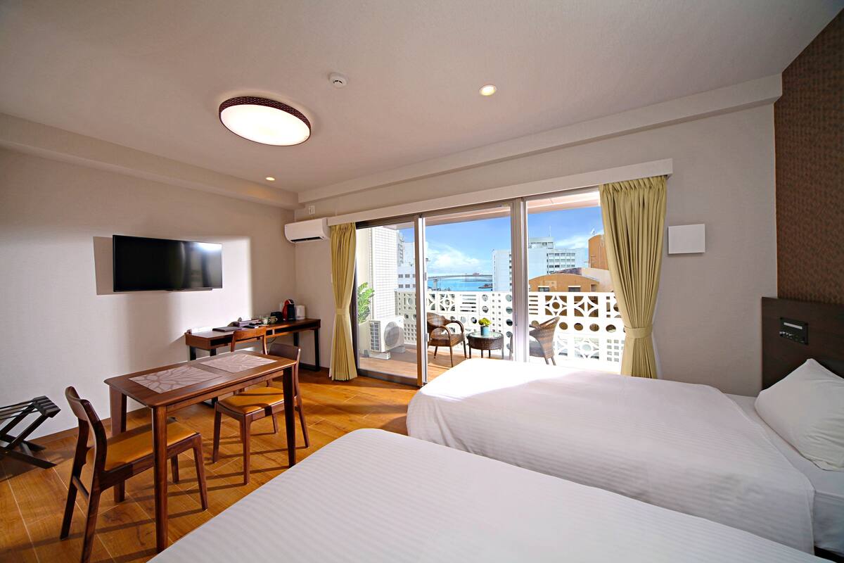 Ein Hotelzimmer mit zwei Betten, einem Esstisch, Stühlen, einem Fernseher und einem Balkon mit Blick auf Gebäude und das Meer.