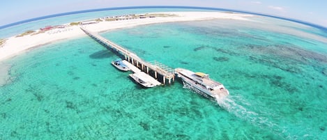 Eine Luftaufnahme eines Strandresorts mit einer Pier, die sich über türkisfarbenes Wasser erstreckt, einem weißen Sandstrand und Bungalows über dem Wasser.
